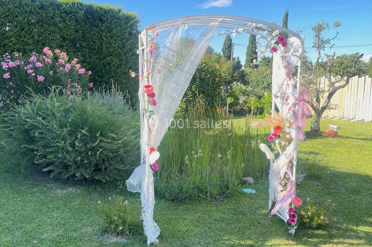 Arche de mariage décorée de fleurs et voilage blanc, installée dans un jardin verdoyant par une journée ensoleillée.