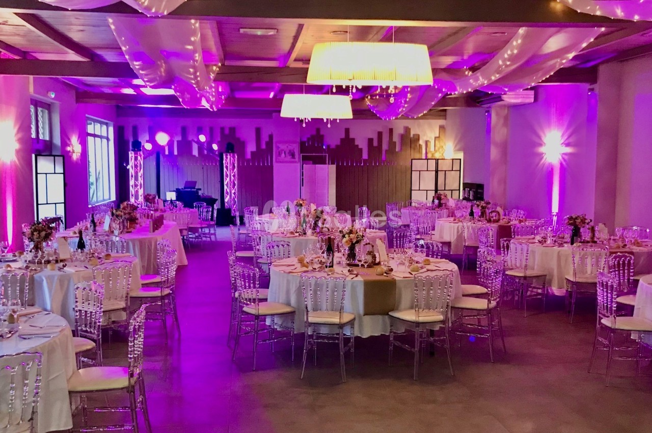 Salle de réception décorée avec des tables rondes dressées, éclairage violet et guirlandes lumineuses au plafond.