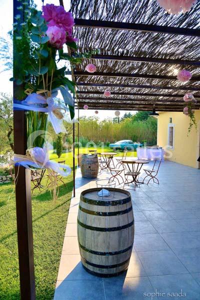 Location salle Saint-Rémy-de-Provence (Bouches-du-Rhône) - Mas de Jonquerolles #19 Terrasse ombragée avec tables en métal, tonneau en bois en premier plan, et jardin verdoyant en arrière-plan.