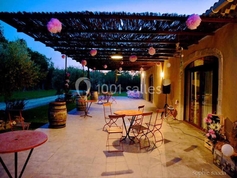 Location salle Saint-Rémy-de-Provence (Bouches-du-Rhône) - Mas de Jonquerolles #9 Terrasse extérieure avec tables et chaises en métal, décorée de lanternes et guirlandes sous une pergola au crépuscule.