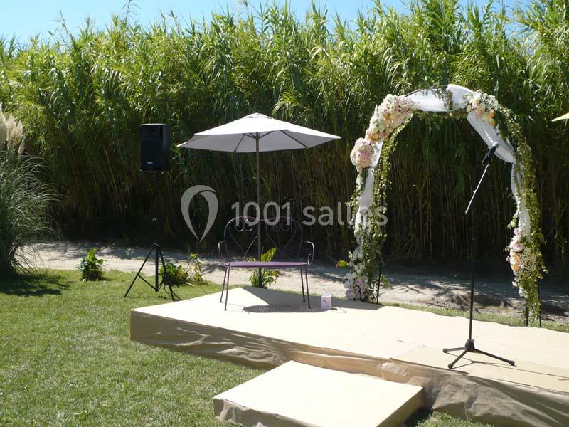 Location salle Saint-Rémy-de-Provence (Bouches-du-Rhône) - Mas de Jonquerolles #23 Estrade en extérieur avec arche fleurie, parasol, banc en métal et micro, entourée de végétation dense.