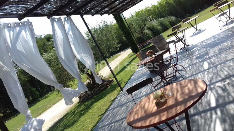 Location salle Saint-Rémy-de-Provence (Bouches-du-Rhône) - Mas de Jonquerolles #4 Terrasse extérieure avec tables, chaises et rideaux blancs flottants, entourée de verdure et d'arbres.