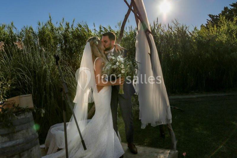 Location salle Saint-Rémy-de-Provence (Bouches-du-Rhône) - Mas de Jonquerolles #11 Un couple en tenue de mariage s'embrasse sous une arche décorée, entouré de verdure et éclairé par le soleil.