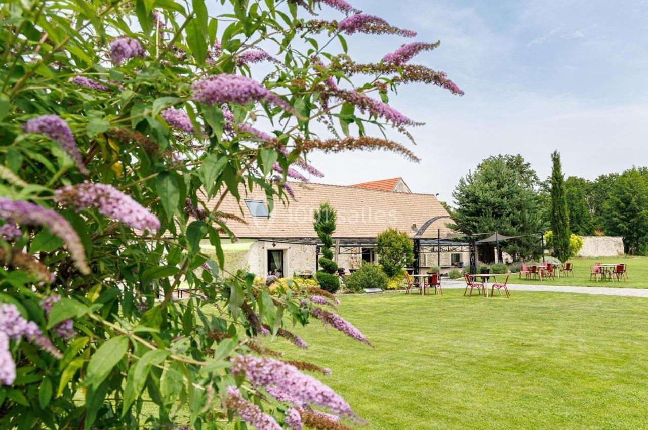 Jardin verdoyant avec pelouse, tables extérieures et bâtiment en pierre, fleurs violettes au premier plan.