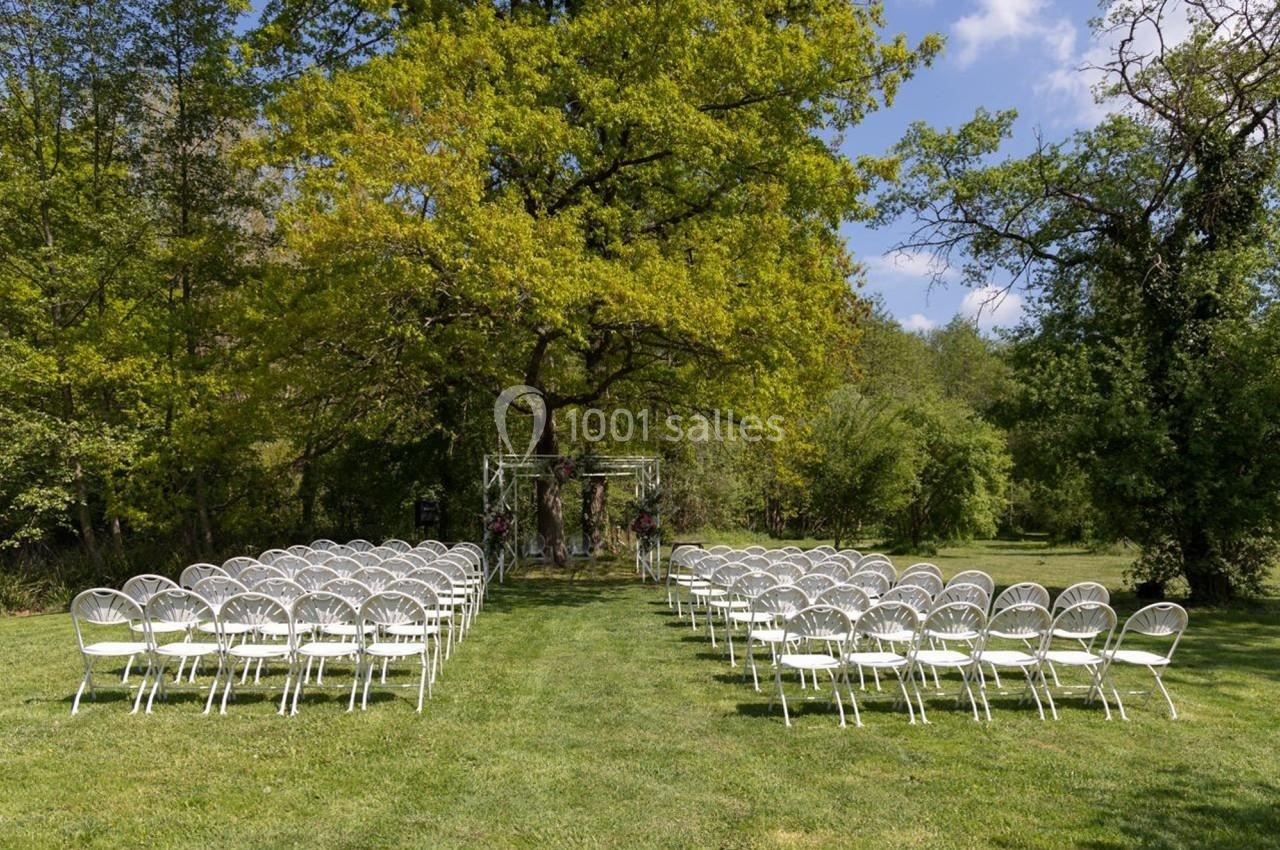 Chaises blanches disposées en rangées sur une pelouse, face à une arche décorée sous un grand arbre, en extérieur.