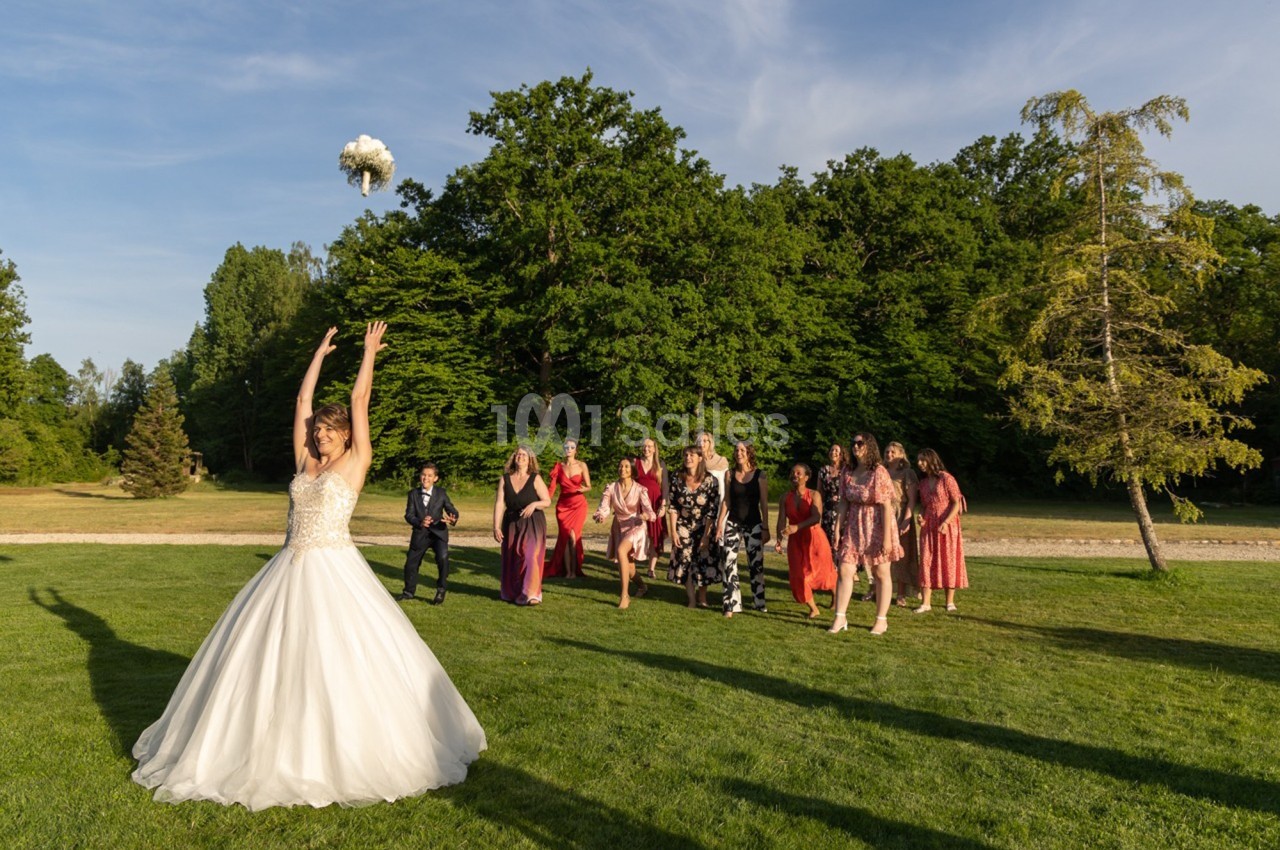 Une mariée lance son bouquet en l'air devant un groupe d'invités prêts à l'attraper, dans un parc verdoyant.