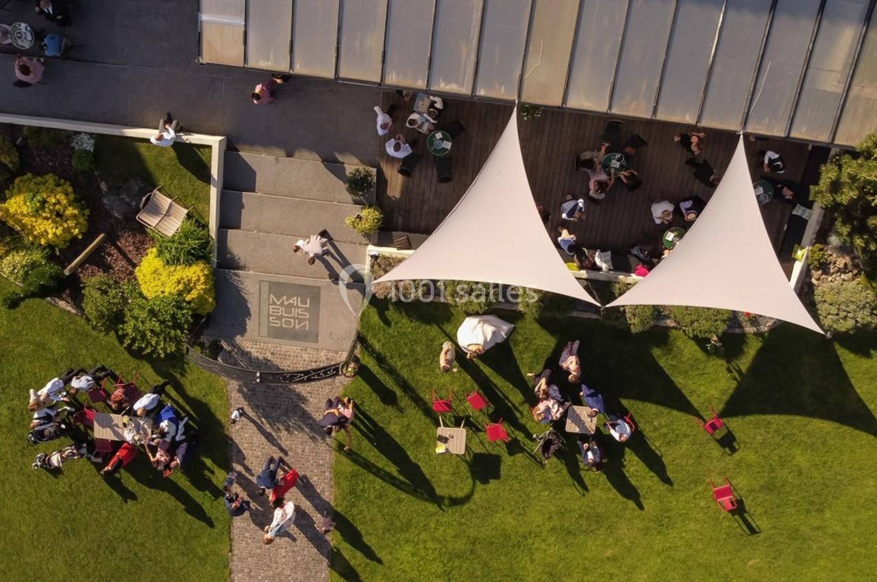 Vue aérienne d'une terrasse avec des voiles d'ombrage, entourée de pelouses et de personnes assises à des tables.