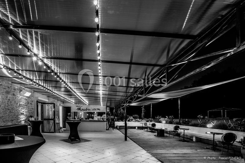 Terrasse couverte éclairée la nuit, avec un bar en arrière-plan et des tables hautes disposées sur le côté.