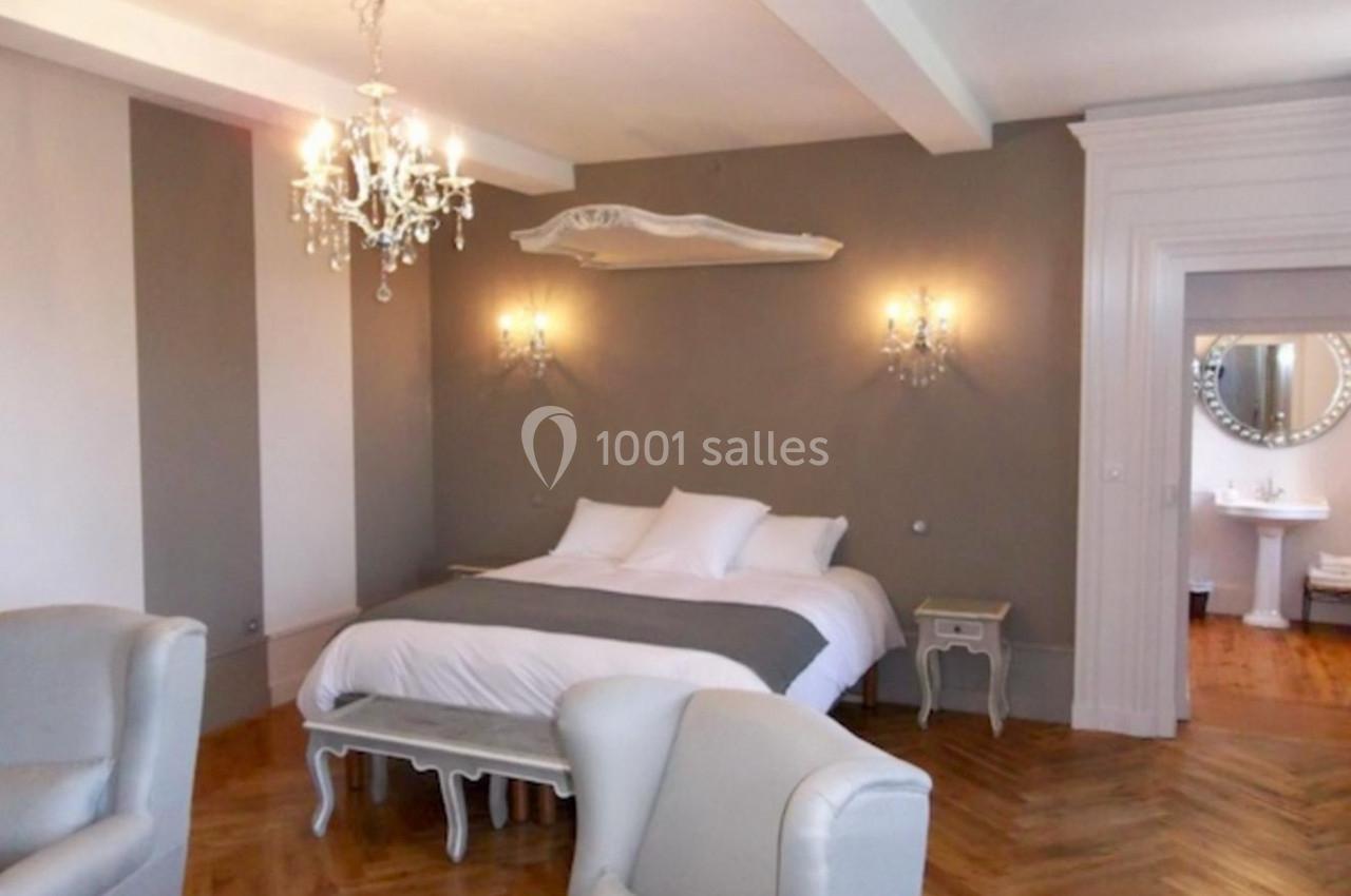 Chambre lumineuse avec lit double, fauteuils, parquet en bois et éclairage mural, vue sur une salle de bain attenante.