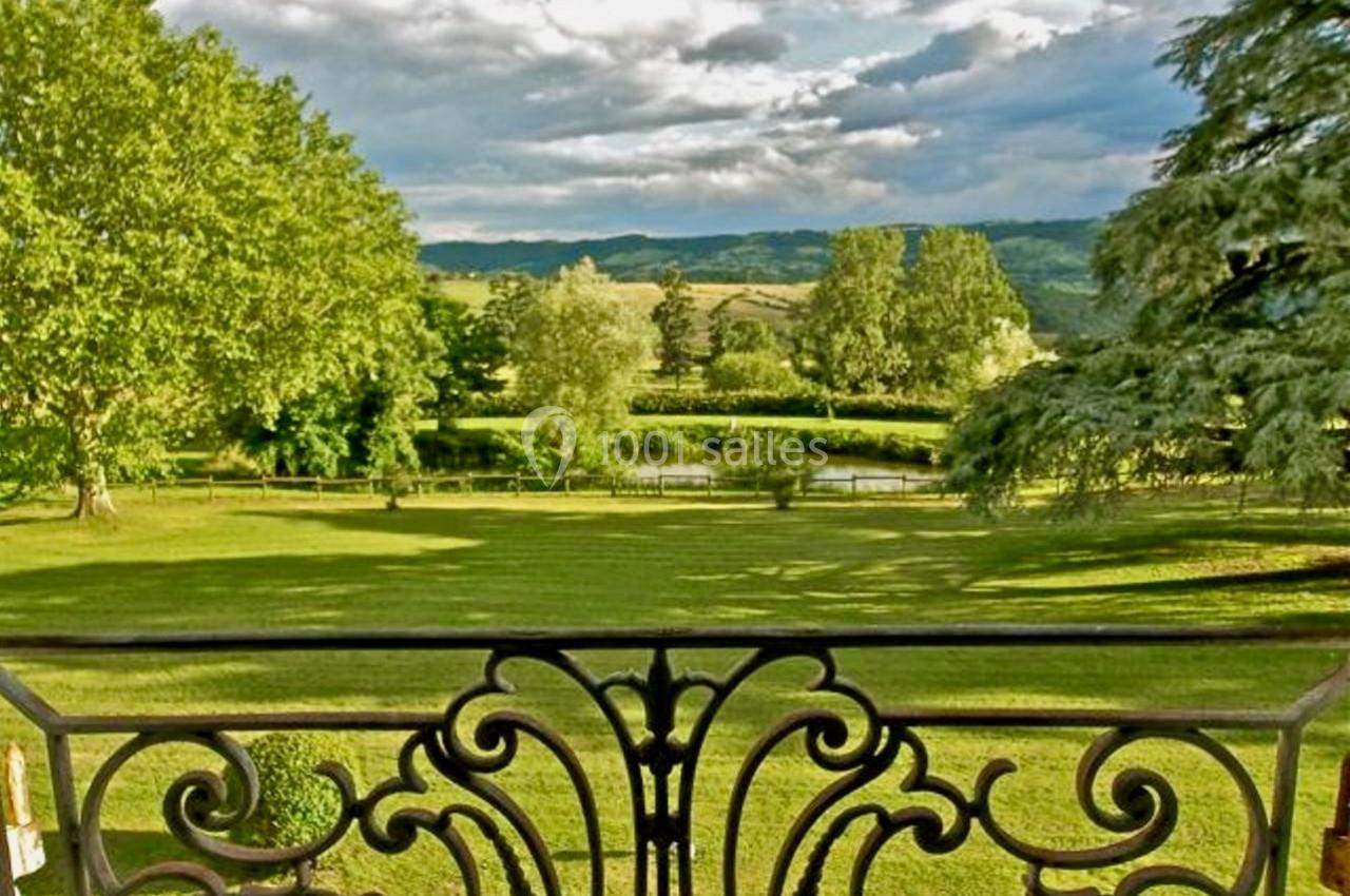 Vue depuis un balcon avec une rambarde en fer forgé sur un parc verdoyant, des arbres et des collines à l'horizon.