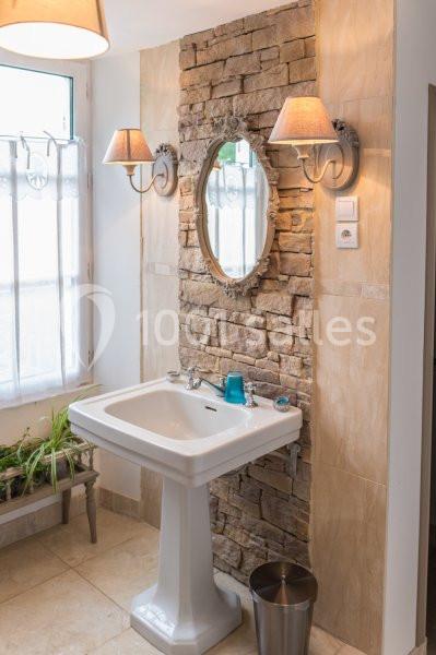 Lavabo blanc sur pied devant un mur en pierres avec miroir ovale, appliques murales et fenêtre à gauche.