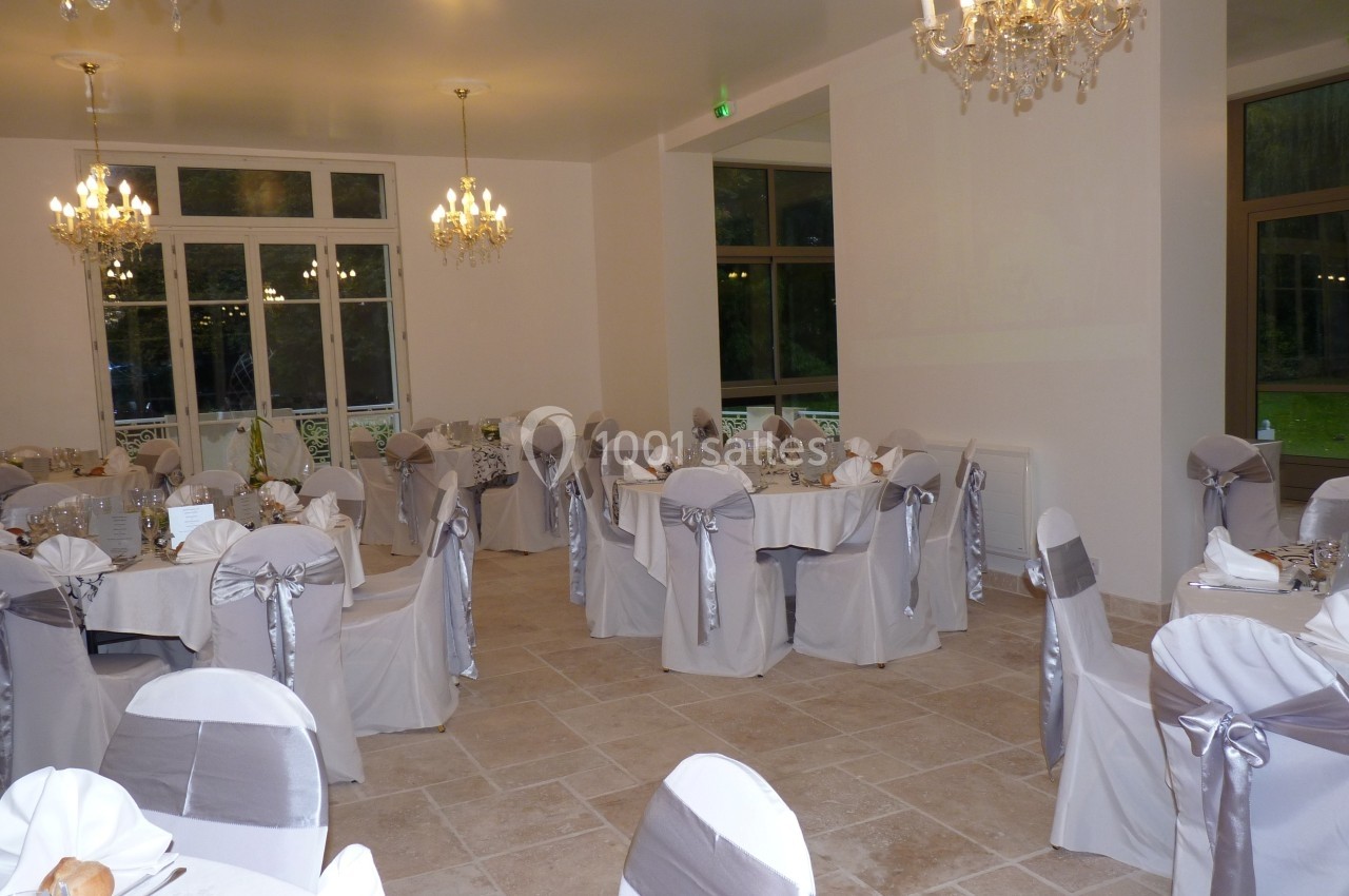 Salle de réception élégante avec tables rondes dressées, chaises décorées de nœuds argentés et lustres suspendus.