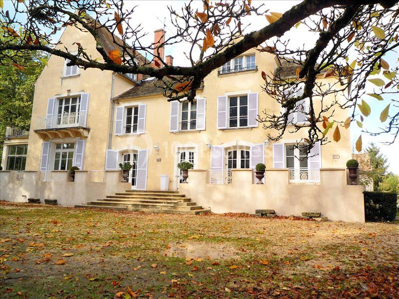 Façade d'une maison ancienne avec volets blancs, entourée de feuilles d'automne et d'arbres.