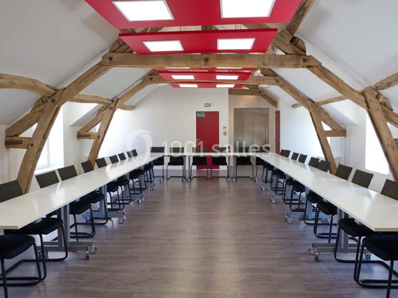 Salle de réunion lumineuse avec poutres apparentes, tables blanches disposées en U et chaises noires.