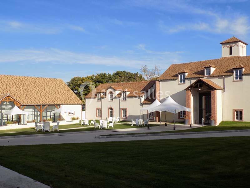 Bâtiments en pierre claire avec toits en tuiles, entourés de pelouses, tables et chaises blanches en extérieur.