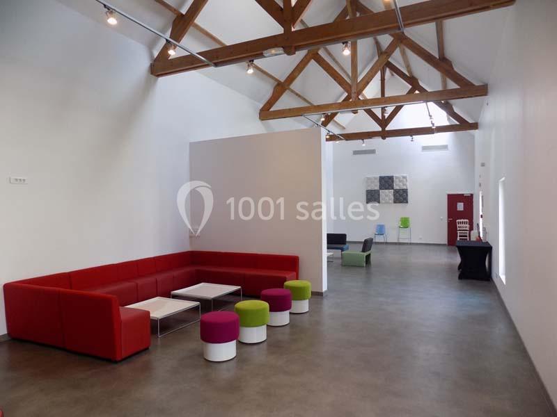 Salle lumineuse avec plafond en poutres apparentes, canapé rouge, tables basses et chaises colorées sur un sol en béton.