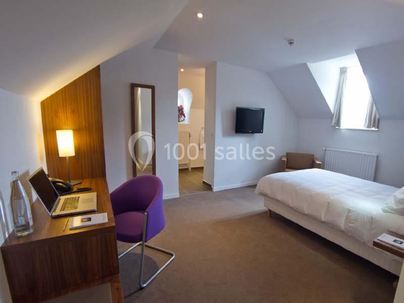 Chambre lumineuse avec lit, bureau en bois, chaise violette, télévision murale et salle de bain attenante.