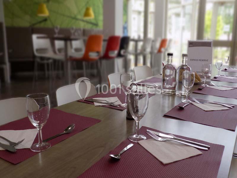 Table dressée avec des sets rouges, verres, couverts et carafes, dans une salle lumineuse avec des chaises colorées.