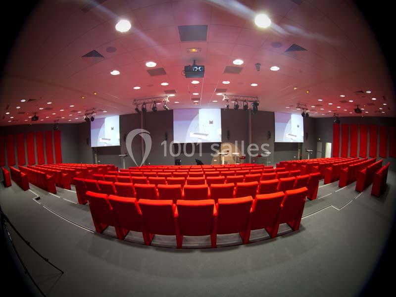 Salle de conférence avec rangées de sièges rouges, écran de projection et éclairage au plafond.