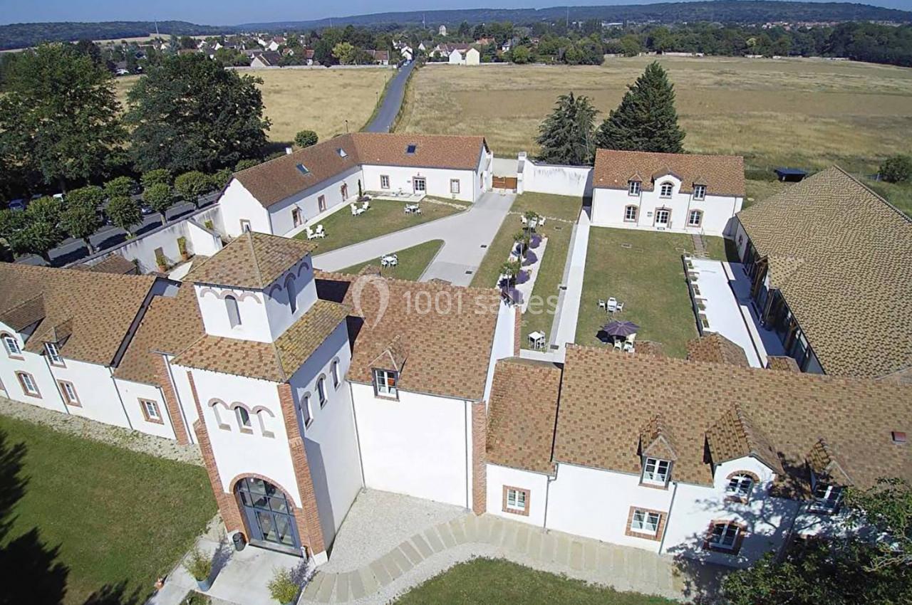 Vue aérienne d'un bâtiment en forme de cour rectangulaire avec des toits en tuiles, entouré de champs et d'arbres.