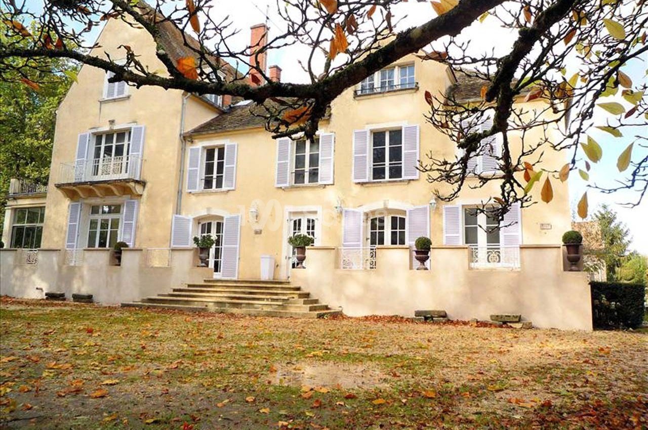 Façade d'une maison ancienne avec volets clairs, entourée d'arbres aux feuilles d'automne tombées au sol.