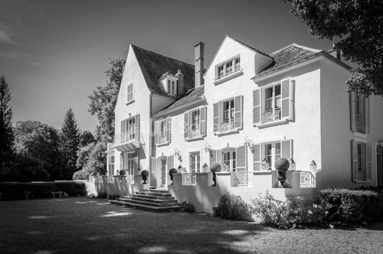Grande maison blanche à deux étages avec volets, entourée d'arbres et d'un jardin, sous un ciel dégagé.