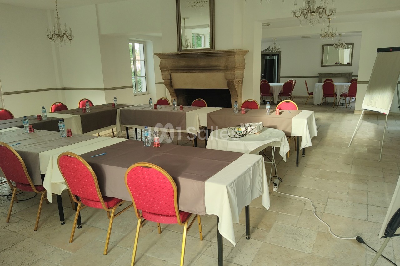 Salle de réunion lumineuse avec tables rectangulaires, chaises rouges, tableau de conférence et cheminée en pierre.