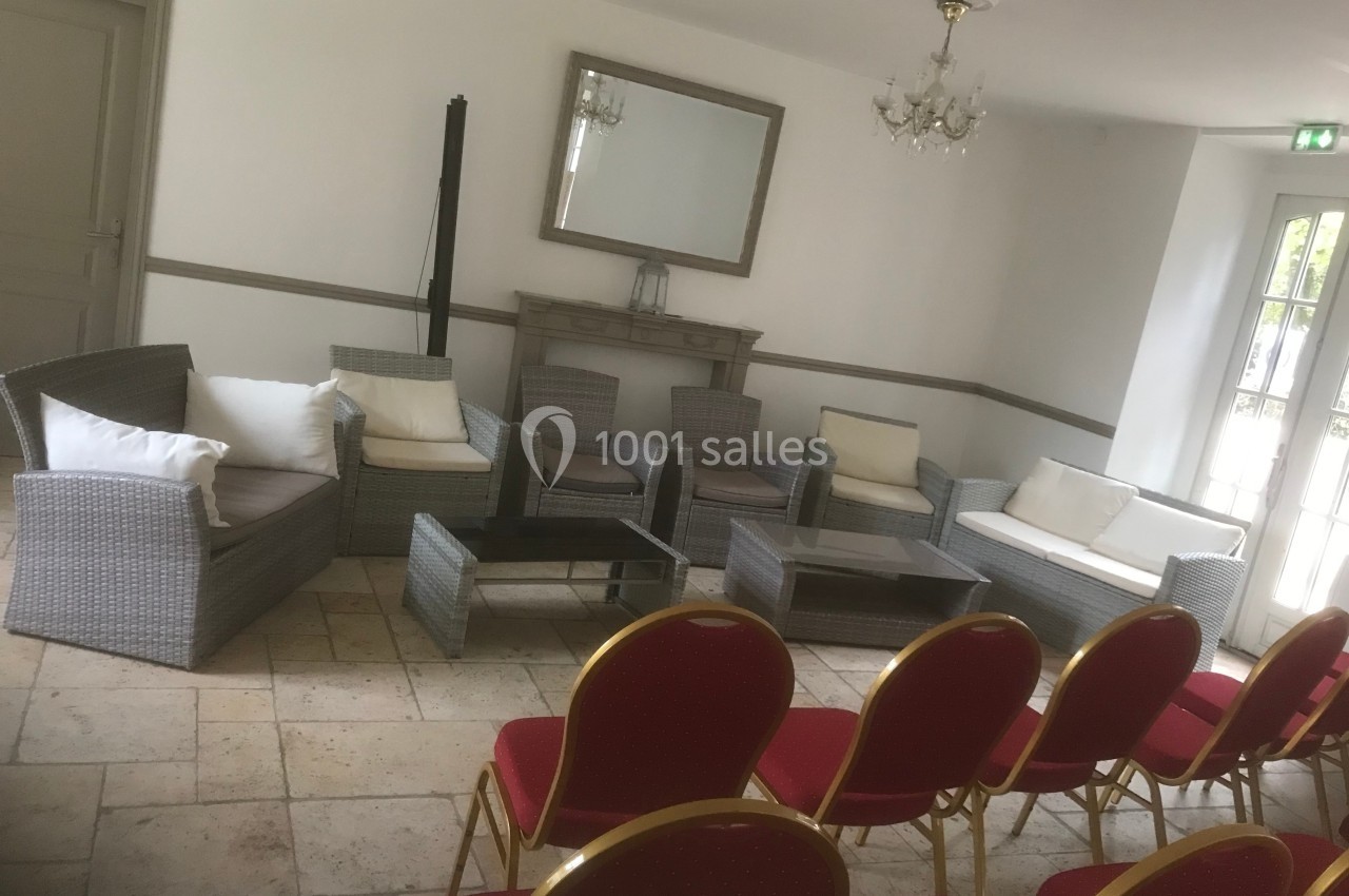 Salle lumineuse avec fauteuils en rotin, tables basses, chaises rouges alignées et cheminée décorative.