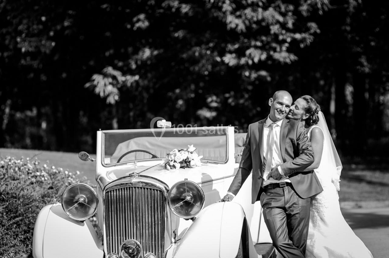Un couple souriant en tenue de mariage pose devant une voiture ancienne blanche dans un cadre extérieur.