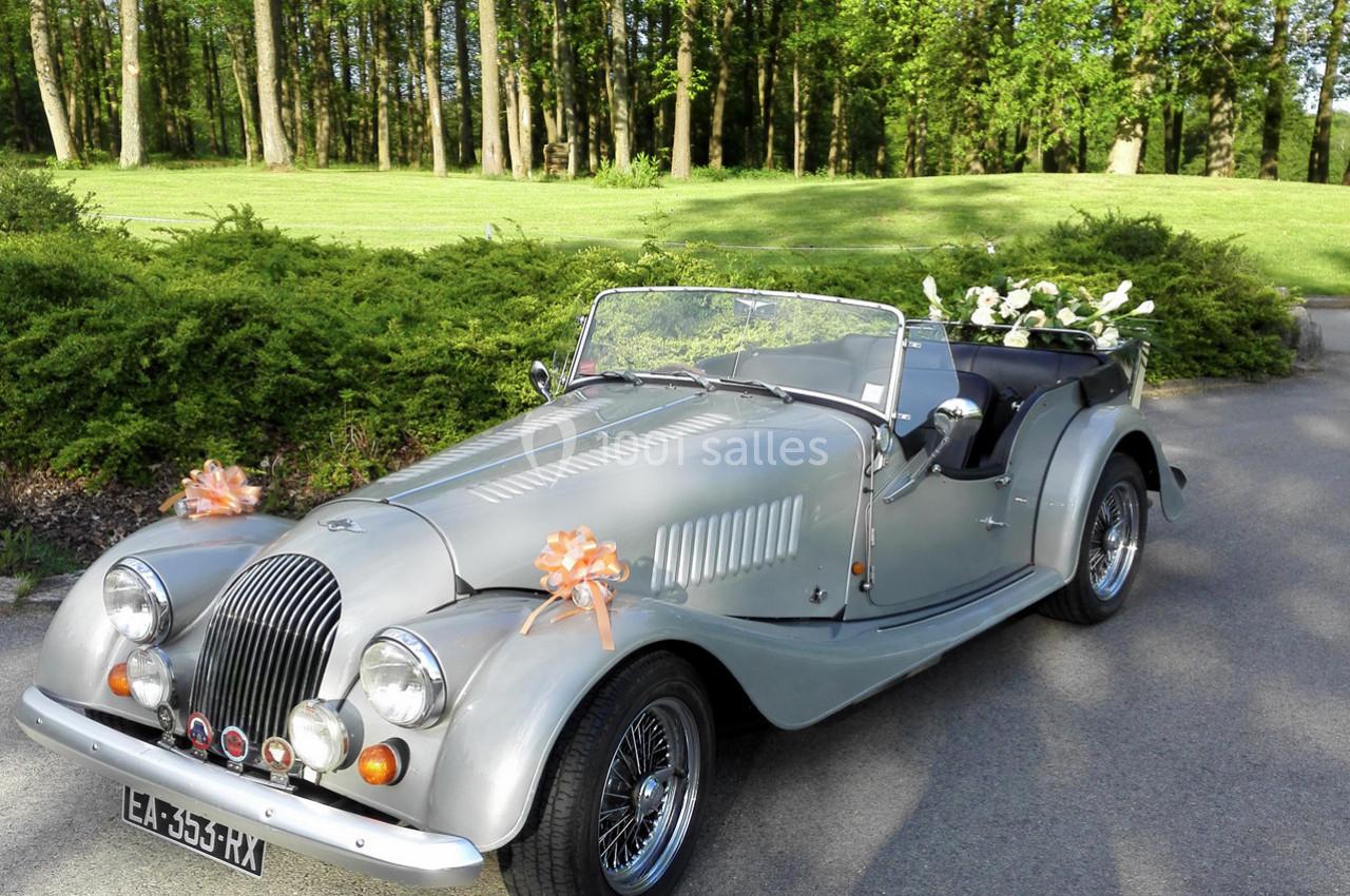 Voiture ancienne décapotable argentée décorée de fleurs et rubans, stationnée sur une allée bordée de verdure.