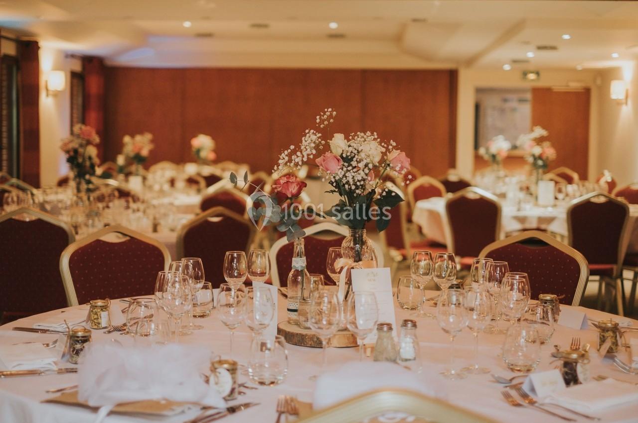 Salle de réception décorée avec des tables rondes, nappes blanches, bouquets de fleurs et verres disposés.