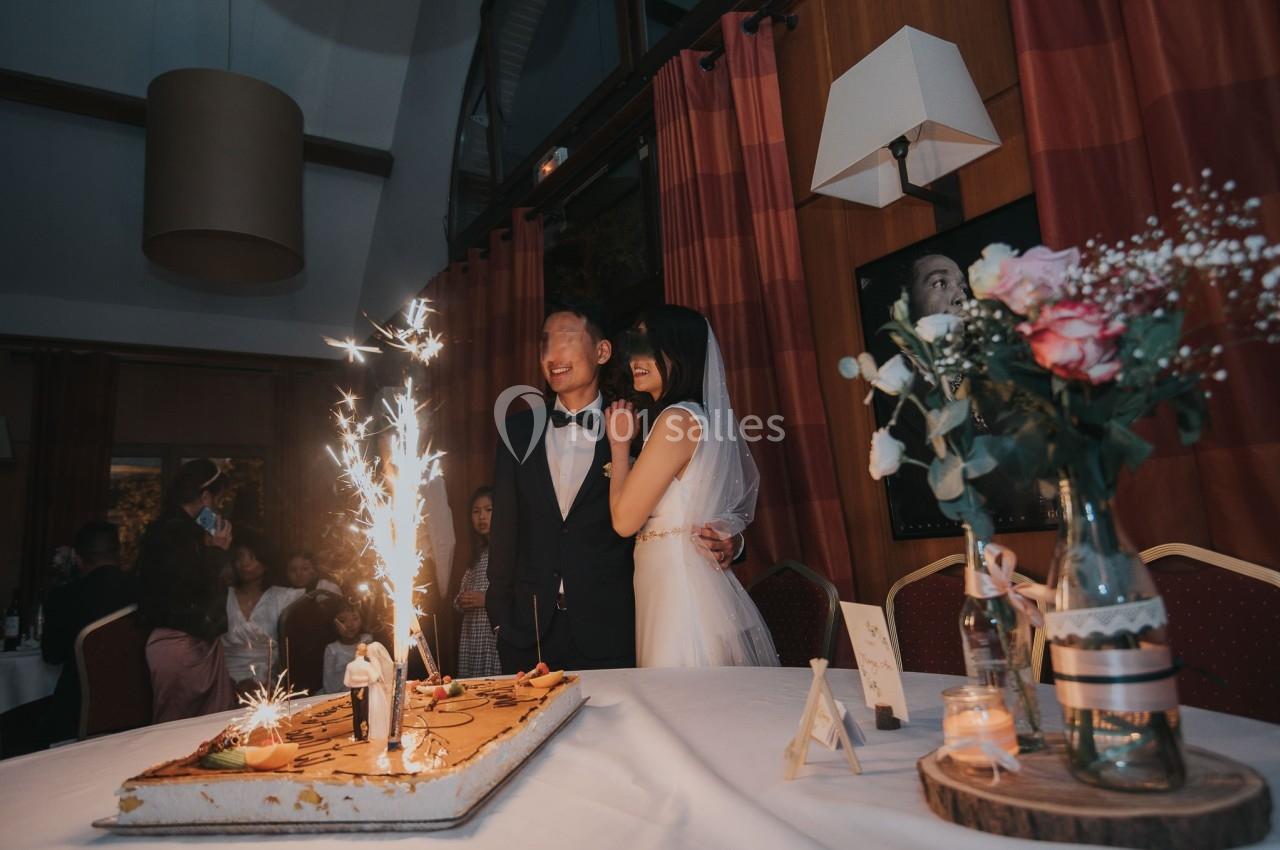 Un couple en tenue de mariage se tient près d'un gâteau décoré de cierges magiques dans une salle de réception.