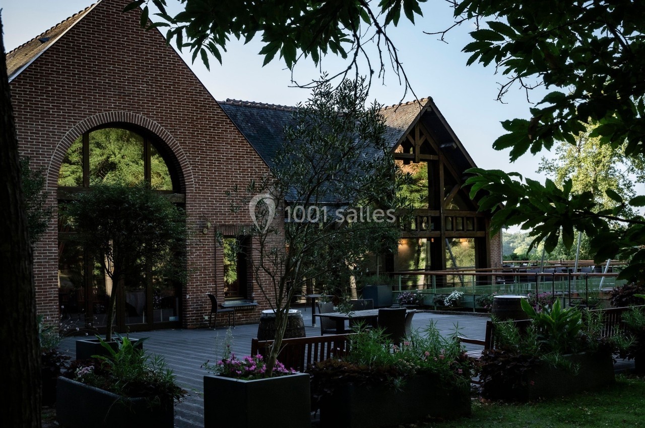 Bâtiment en briques avec de grandes baies vitrées, entouré de végétation et d'une terrasse aménagée.