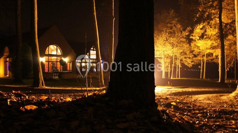 Maison éclairée entourée d'arbres dans une ambiance nocturne, avec des feuilles au sol et une lumière chaude.