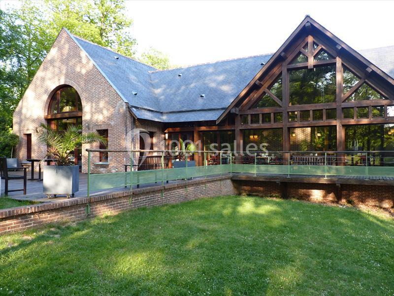 Maison en briques avec grandes baies vitrées, terrasse en bois, rampe en verre et jardin verdoyant.