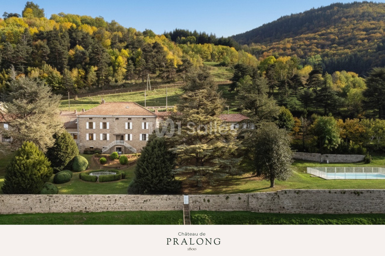 Vue d'un château en pierre entouré de jardins, d'arbres et de collines boisées sous un ciel dégagé.
