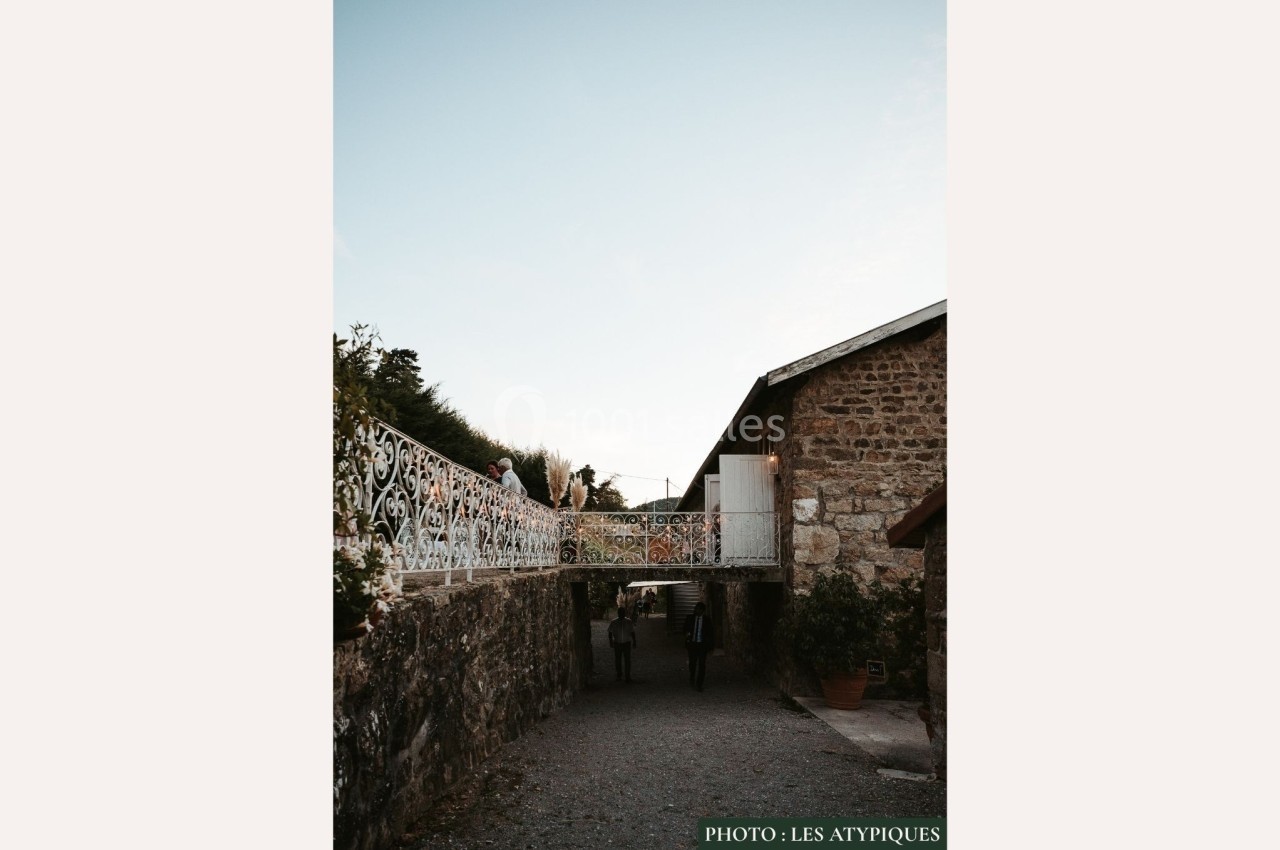 Passage en pierre avec une passerelle métallique décorative, menant à des bâtiments sous un ciel dégagé.