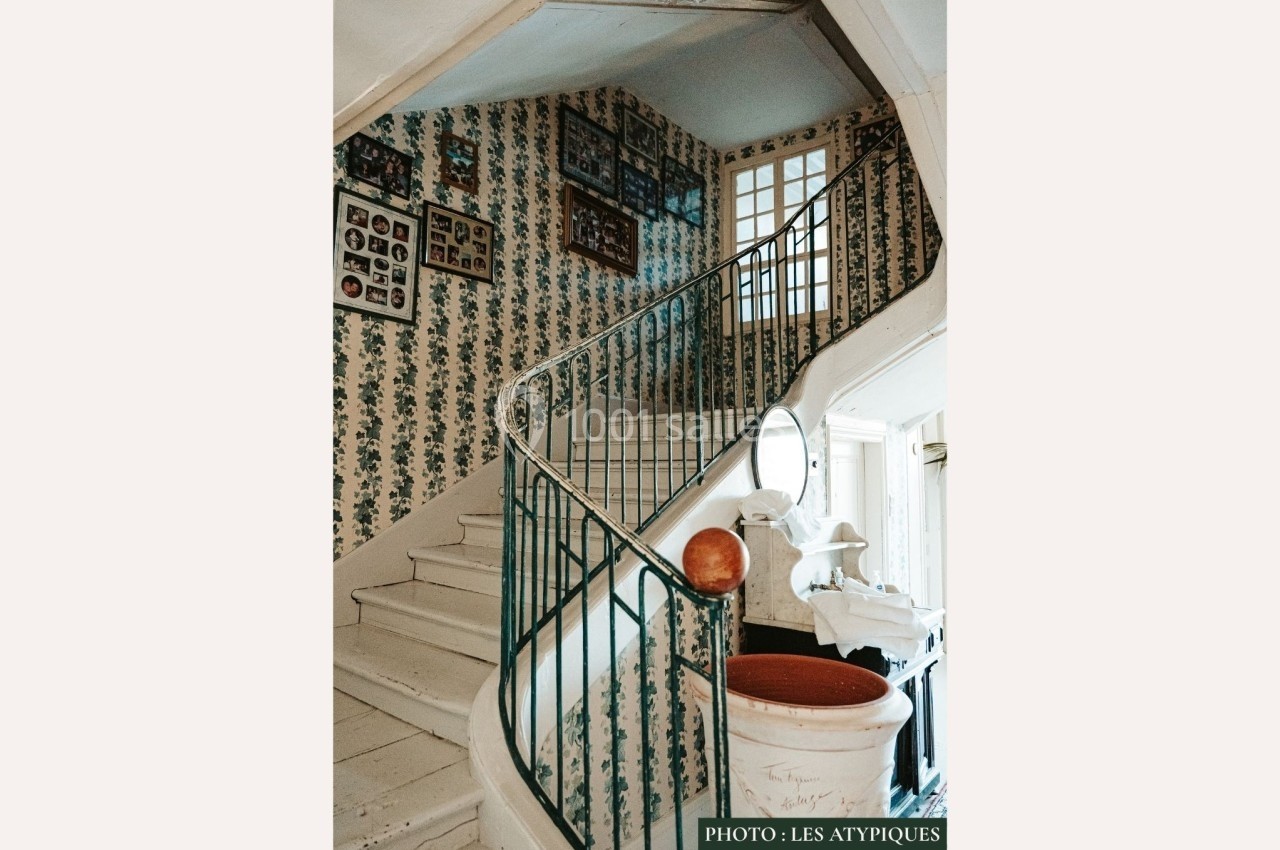 Escalier ancien en bois peint en blanc, entouré de murs tapissés de motifs floraux et décoré de cadres et objets vintage.