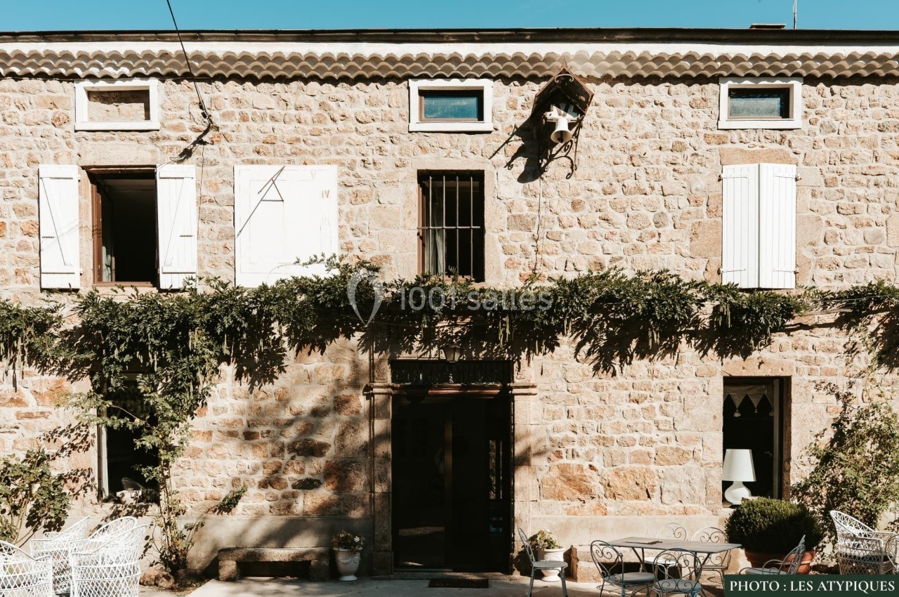 Façade en pierre d'une maison avec volets blancs, vigne grimpante et mobilier de jardin sur une terrasse.