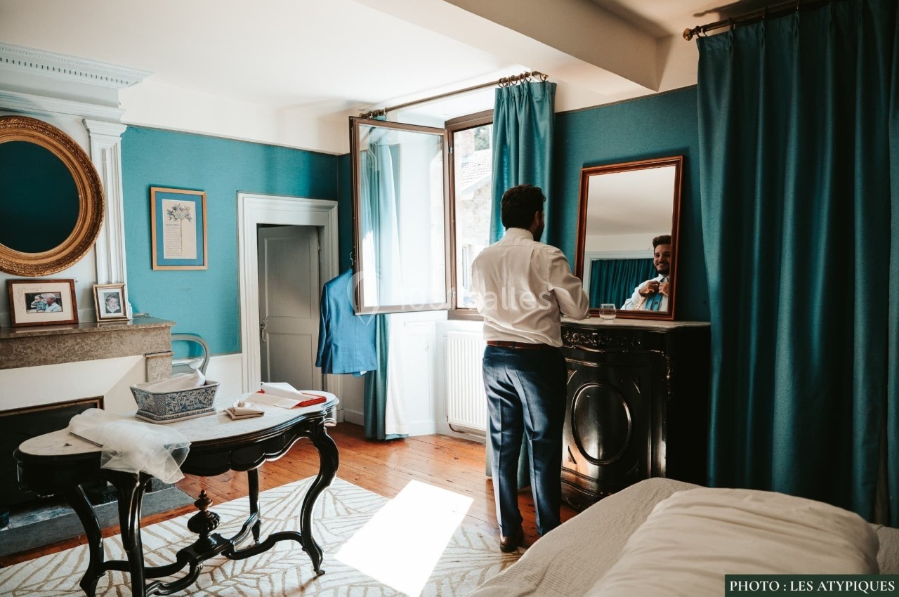 Un homme en chemise se prépare devant un miroir dans une chambre lumineuse aux murs bleus et rideaux assortis.