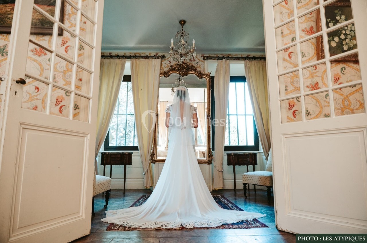 Une mariée en robe blanche se tient devant un grand miroir dans une pièce élégante avec des portes vitrées ouvertes.