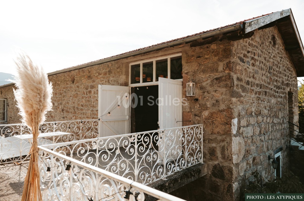 Façade en pierre d'une maison avec balcon en fer forgé blanc, portes ouvertes et décoration de pampas.