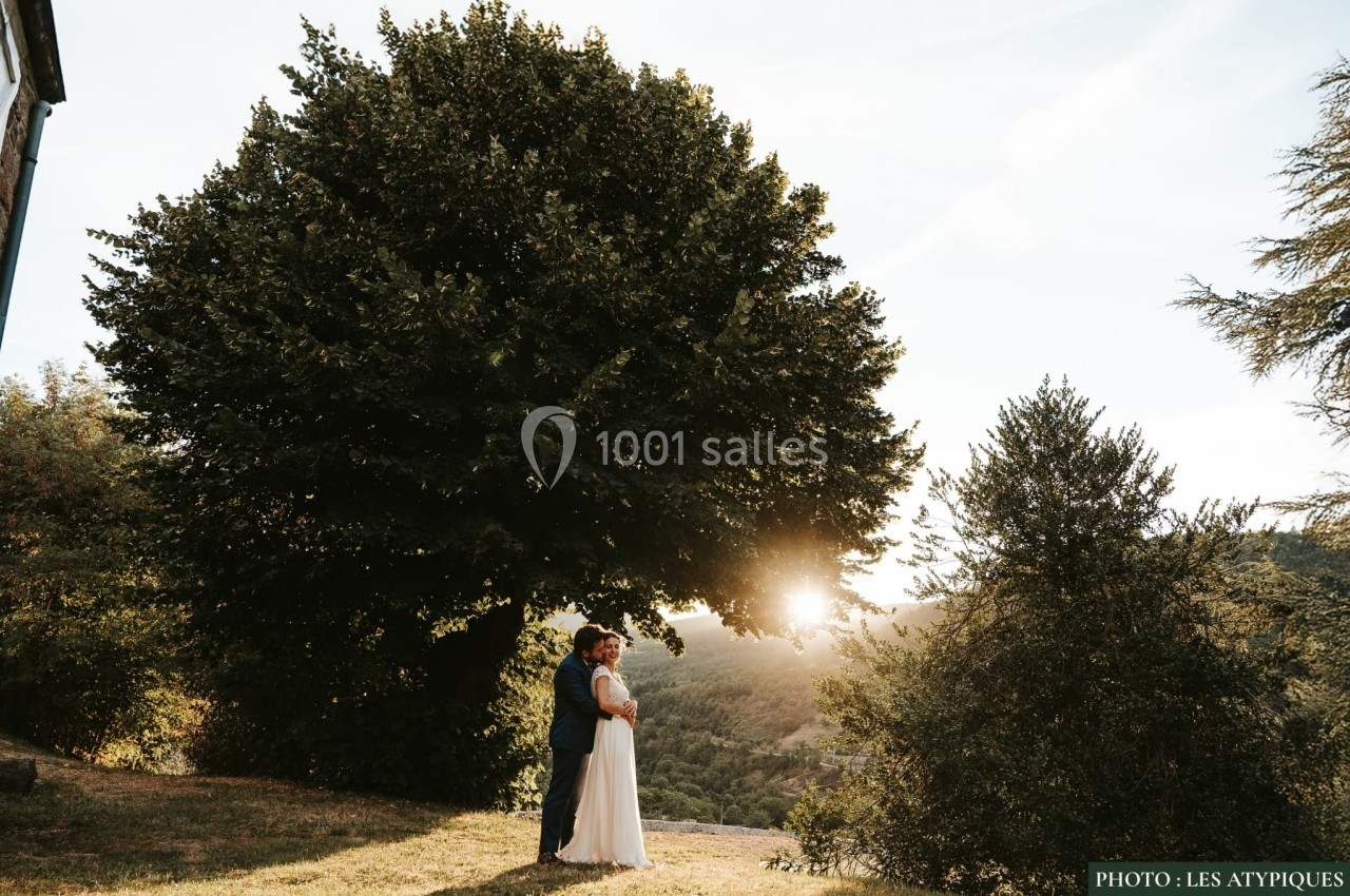 Un couple debout sous un grand arbre au coucher du soleil, entouré de verdure et de collines.