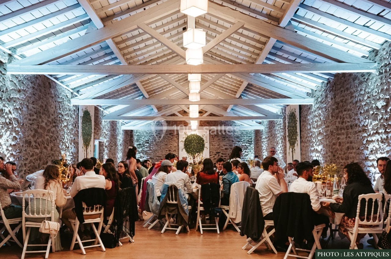 Salle de réception en pierre avec des invités assis à des tables décorées, éclairée par des suspensions lumineuses.