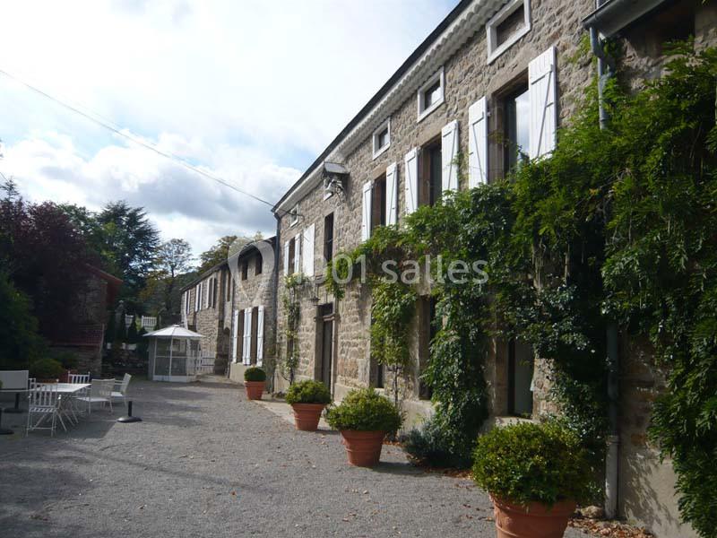 Façade en pierre d'une maison avec volets blancs, plantes grimpantes et terrasse aménagée avec des pots et des chaises.