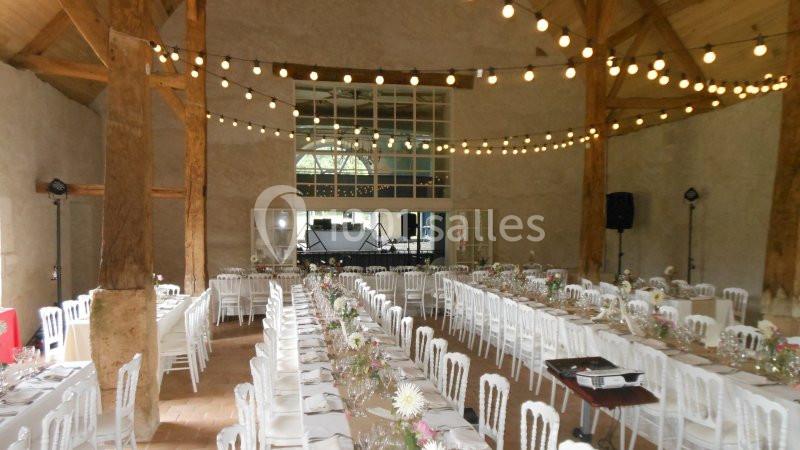Salle de réception avec de longues tables décorées, des chaises blanches et des guirlandes lumineuses suspendues.