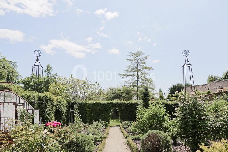 Allée centrale bordée de rosiers et arbustes dans un jardin fleuri, sous un ciel bleu parsemé de nuages.