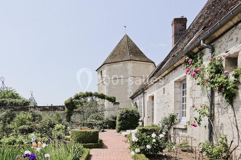 Jardin fleuri avec allée en briques, bordé de rosiers, menant à une tour en pierre sous un ciel dégagé.