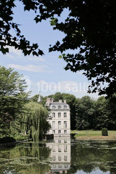 Vue d'un bâtiment ancien entouré de verdure, reflété dans un étang calme sous un ciel partiellement dégagé.