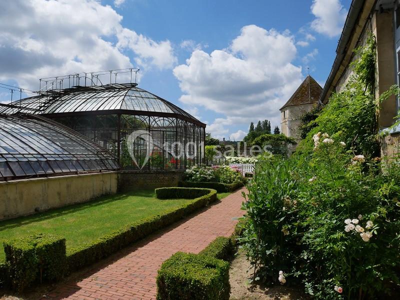 Chemin pavé bordé de haies et de fleurs menant à une serre vitrée dans un jardin sous un ciel partiellement nuageux.